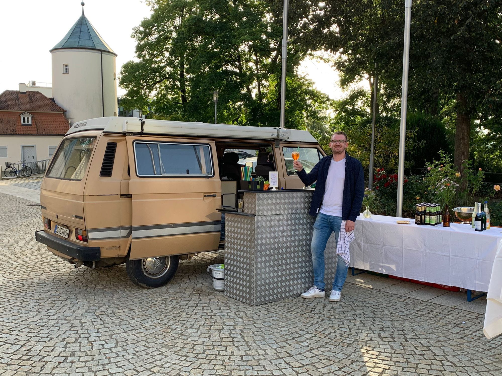 Moritz Bayer steht mit einem Aperol vor seiner mobilen Bar. Der alte VW Bus gehört zur Bar dazu